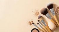 Professional makeup brushes and cosmetics arranged on neutral beige background with scattered powder creating elegant beauty flatlay composition for skincare routine.