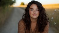 Young woman with long dark wavy hair and natural makeup smiling warmly at sunset outdoors. Soft golden hour lighting creates dreamy atmosphere with bokeh bubbles.
