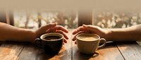 Two hands reaching for coffee cups on wooden table in warm morning light creating intimate moment for cafe and relationship concepts.