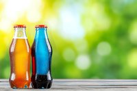 Two refreshing soda bottles with condensation drops on wooden surface against blurred green natural background, orange and blue colored drinks.