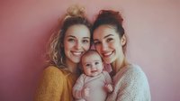 Two young Caucasian women with messy buns holding adorable baby against soft pink background. Happy mothers or sisters smiling warmly in cozy sweaters for family lifestyle.