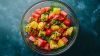 Fresh tropical fruit salad with watermelon, pineapple, strawberries and mint leaves in glass bowl on dark turquoise background, summer refreshment.