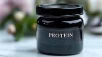 Black protein powder container with label displayed on clean surface with blurred flowers and wellness items in background for nutritional supplement marketing.