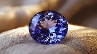 Oval cut tanzanite gemstone with deep violet-blue color displaying brilliant facets and exceptional clarity on natural linen fabric background. Luxury jewelry element.