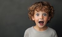 Excited young boy with curly ginger hair and wide open mouth expressing pure joy and surprise against dark background, wearing gray t-shirt. For advertising and emotion concepts.