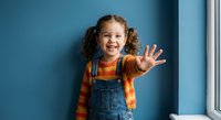 Happy young girl with curly hair waving hand wearing denim overalls and striped shirt against blue wall background for children lifestyle concepts.