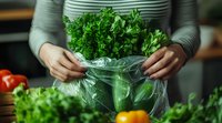 Fresh parsley bunch in plastic bag held by hands in striped sweater, storing herbs in kitchen with vegetables in background, food preparation concept.