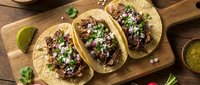 Three corn tortillas filled with shredded beef, topped with fresh cilantro, onions and lime wedges on wooden board, ready for serving or restaurant menu use.