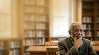 Senior African American male scholar in glasses wearing olive cardigan, seated at wooden desk in library with bookshelves, contemplative pose for education and academic content.