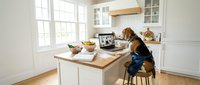 Young woman with long brown hair working on laptop at modern kitchen island counter with white cabinets and natural lighting for remote work lifestyle.