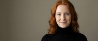 Professional redhead woman in black turtleneck sweater smiling confidently against neutral background for business and corporate marketing materials.