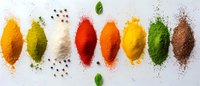Colorful spice powders in row with peppercorns and basil leaves on white background, overhead view. Food seasoning collection showing vibrant cooking ingredients.