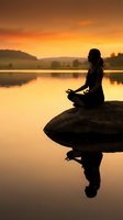 Woman meditating in lotus position on rock at sunrise by calm lake, silhouetted against golden sky with mountain backdrop, reflecting on water for wellness and mindfulness concepts.