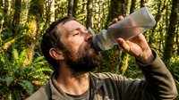 Middle-aged Caucasian male hiker drinking water from bottle in lush forest environment. Man refreshing during outdoor adventure among green trees and natural woodland setting.
