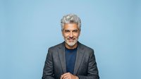 Mature man with gray hair and mustache wearing dark blazer and blue shirt against blue background, suitable for corporate portraits and business headshots.