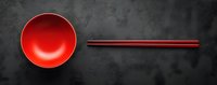Red ceramic bowl and traditional chopsticks arranged on dark stone background, minimalist Asian dining composition for restaurant menu and food presentation.