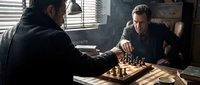 Two middle-aged Caucasian men in dark clothing playing chess in dimly lit room with venetian blinds casting dramatic shadows across wooden table.