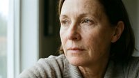 Middle-aged woman with fair skin and freckles, wearing a neutral beige sweater, gazing thoughtfully toward a window with natural light. Suitable for healthcare, wellness, and lifestyle editorial use.