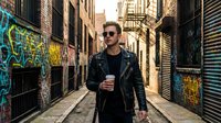 Young Caucasian man in leather jacket and sunglasses walking through urban alley with colorful graffiti street art, holding coffee cup in city environment.