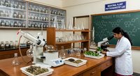 Young Asian female scientist in white lab coat conducting research with microscope in vintage chemistry laboratory with glass bottles and chalkboard.