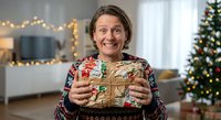 Middle-aged Caucasian woman holding wrapped Christmas gift with festive sweater in decorated living room with tree and warm lights during holiday season.