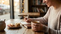 Woman enjoying morning coffee while writing in cozy cafe with warm lighting and bookshelves creating peaceful atmosphere for journaling and relaxation.