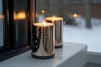 Glowing candles in metallic holders on windowsill against snowy winter background create cozy atmospheric mood with bokeh light effects and raindrops on window glass.