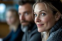 Middle-aged woman with blonde hair smiling at camera in indoor setting with blurred colleagues in background. Suitable for corporate, business, teamwork, and professional workplace concepts.