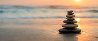 Zen stone stack on beach at sunset with ocean waves and golden light creating peaceful meditation atmosphere for wellness and mindfulness practices.