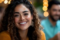 Young Latin woman with curly hair and bright smile wearing yellow sweater against blurred bokeh lights background, natural beauty portrait.