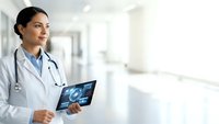 Female Hispanic doctor holding digital tablet with medical interface in modern hospital corridor for healthcare technology presentations.