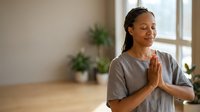 Middle-aged woman of South Asian descent practicing yoga indoors, hands in prayer position at chest, wearing gray t-shirt in bright studio with plants and windows, mindfulness and wellness concept.