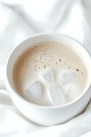 Creamy beige drink with ice cubes in white ceramic cup against light fabric background, featuring delicate foam bubbles and soft minimalist composition.