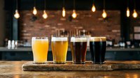 Four beer glasses with different shades arranged on wooden board in pub setting with hanging lights and brick wall background for brewery, bar, or beverage industry use.