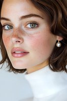 Close-up portrait of young woman with natural freckles, brown hair and blue eyes wearing white turtleneck and pearl earring against light background.