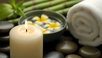 Spa wellness setup with burning candle, rolled towel, floating flowers in water bowl and smooth stones for relaxation and massage therapy.