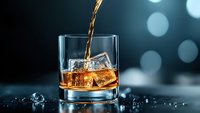 Pouring golden whiskey into rocks glass with ice cubes on dark background with bokeh lights, splashing liquid creates dynamic motion effect for beverage photography.