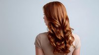 Young woman with long wavy copper red hair shown from behind against light gray background, demonstrating professional hairstyling and hair coloring result.