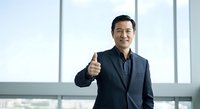 Mature Asian male professional in dark blazer giving thumbs up gesture in modern office with floor-to-ceiling windows, expressing approval, success, and positive business attitude.
