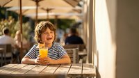 Happy young boy with curly hair enjoying fresh orange juice at outdoor cafe table during sunny day, smiling at camera in casual striped shirt.