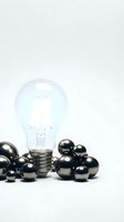 Clear incandescent light bulb with glowing filament surrounded by shiny black spheres on white background, representing innovation and creative ideas for business concepts.