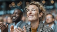 Middle-aged Caucasian woman with curly hair smiling joyfully at corporate event or conference. Professional audience applauding in background with warm lighting.