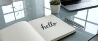 Open notebook with handwritten hello on a modern glass desk beside a pen, succulent plant, and laptop in a bright workspace.