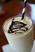 Creamy vanilla milkshake with chocolate syrup drizzle and striped straw in clear glass, served on wooden table for refreshing summer treat.