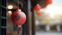 Traditional red paper lantern with ornate pattern and tassel hanging outdoors against blurred background with warm evening light and additional lanterns.