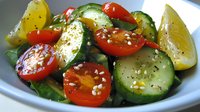 Fresh summer salad with sliced cucumber, cherry tomatoes, and lemon wedges garnished with sesame seeds and herbs in white ceramic bowl, close up view.