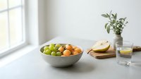 Assorted fresh fruits in metallic bowl on white kitchen counter with bread, water glass and potted plant for healthy eating and home decor.