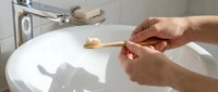 Hands holding a bamboo toothbrush with white toothpaste over a clean bathroom sink, promoting eco-friendly oral hygiene and sustainable daily routine.