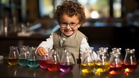Young curly-haired boy in glasses and vest smiling while conducting colorful science experiment with glass beakers containing rainbow liquid solutions in classroom.