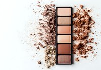 Crushed powder eyeshadow palette in neutral earth tones with scattered powder texture on white background, makeup product photography for beauty industry.